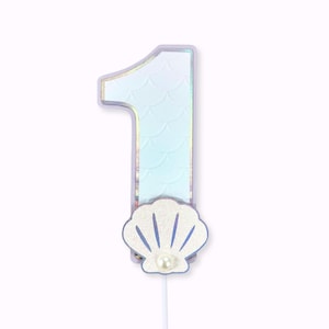 May include: A number one birthday cake topper with a mermaid theme. The number is light blue with a scale pattern and a shimmering iridescent border. A white seashell with a pearl is at the base of the number.