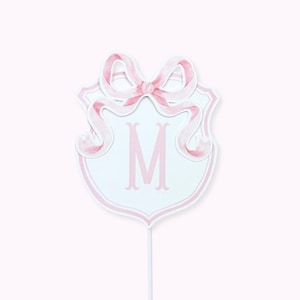 May include: A pink and white decorative shield-shaped topper with a pink bow and the letter "M". The topper is on a white stick, perfect for cakes or other celebratory items. The design features a watercolor style.