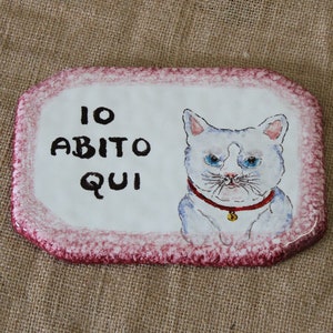 May include: White ceramic tile with a pink border. The tile features a hand-painted image of a white cat with blue eyes and a red collar with a bell. The tile also has the text "IO ABITO QUI" written in black.