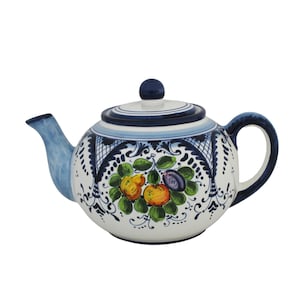 Può includere: Una teiera in ceramica bianca con manico, beccuccio e coperchio blu. La teiera è decorata con un motivo floreale con frutta gialla e arancione, foglie verdi e un bordo decorativo blu e bianco. Il coperchio ha un piccolo pomello blu.