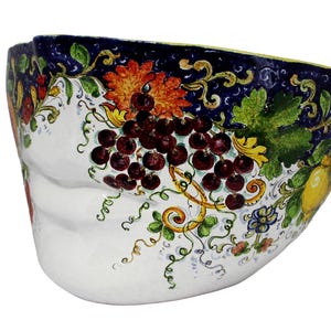 以下が含まれることがあります： 暗い赤色のブドウ、黄色のレモン、オレンジ色の花を手描きでデザインした白いセラミックボウル。縁には装飾模様の濃い青色の縁取りがあります。ボウルはユニークで非対称な形状をしています。