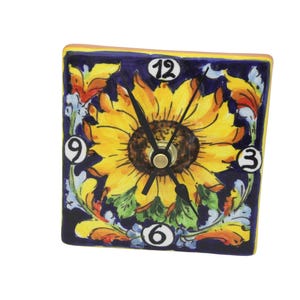 May include: Square ceramic clock with a sunflower design. The clock face features a yellow sunflower with a brown center, set against a dark blue background. The clock has black hands and white numerals. The clock is trimmed with a yellow border.