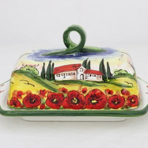 May include: A ceramic butter dish with a green handle and a painted scene of a house with red tile roof, green trees, and red poppies on a yellow field.