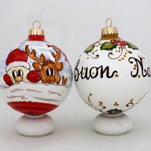 Può includere: Due addobbi natalizi in ceramica dipinti a mano. Un addobbo presenta Babbo Natale e una renna che spuntano da un paesaggio innevato. L'altro addobbo è bianco con dettagli dorati e le parole "Buon Natale" scritte in nero.