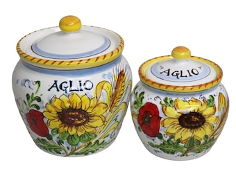 Op de afbeelding: Twee keramische potten met deksels. De potten zijn wit met gele, blauwe en rode bloemmotieven, waaronder zonnebloemen en klaprozen. Het woord "AGLIO" staat op de potten.