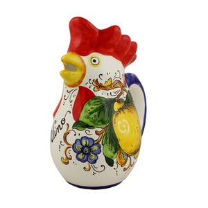 Puede incluir: Jarra de cerámica con forma de gallo, con cresta roja, pico amarillo y cuerpo blanco. La jarra está decorada con un diseño de limón y flores, con la palabra "vino" escrita en el lateral. El asa es azul.