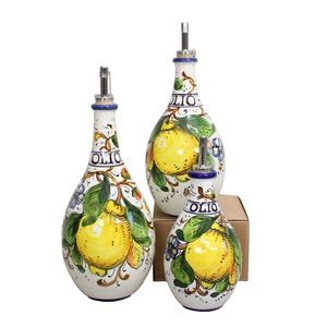 Può includere: Set di tre bottiglie in ceramica per olio d'oliva con base bianca, con limoni gialli dipinti a mano, foglie verdi e fiori blu. Ogni bottiglia ha un beccuccio argentato e la parola "OLIO" è scritta sul collo.