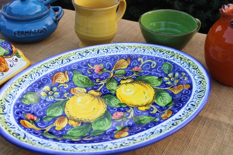 Puede incluir: Una fuente ovalada de cer&aacute;mica con fondo azul y dise&ntilde;o de limones amarillos y verdes. La fuente est&aacute; rodeada de otras piezas de cer&aacute;mica coloridas, incluyendo un cuenco azul con la palabra "FORMAGGIO" y una taza amarilla.