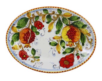 May include: Oval ceramic serving platter with a white base, featuring hand-painted pomegranates, red flowers, and green leaves. The rim has a blue border with a yellow and orange trim. A decorative piece for serving food.