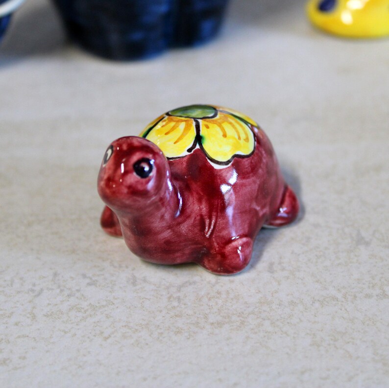 May include: A small, ceramic turtle figurine. The turtle is a deep burgundy color with a yellow and green floral design on its shell. The figurine has a glossy finish and is set against a neutral background.