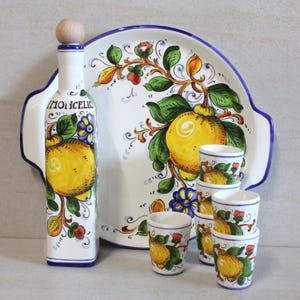 Può includere: Un vassoio da portata in ceramica bianca con un bordo blu e un design a limone. Il vassoio è accompagnato da una bottiglia e sei bicchieri da shot, tutti con un design a limone simile.