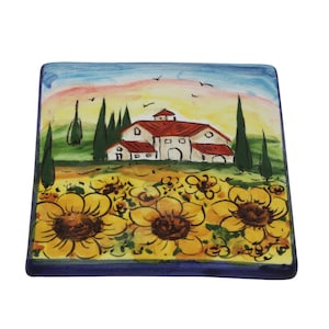May include: Square ceramic coaster with a hand-painted design. The artwork depicts a rural scene with a farmhouse, sunflowers, and trees under a colorful sky. The coaster has a dark blue border.