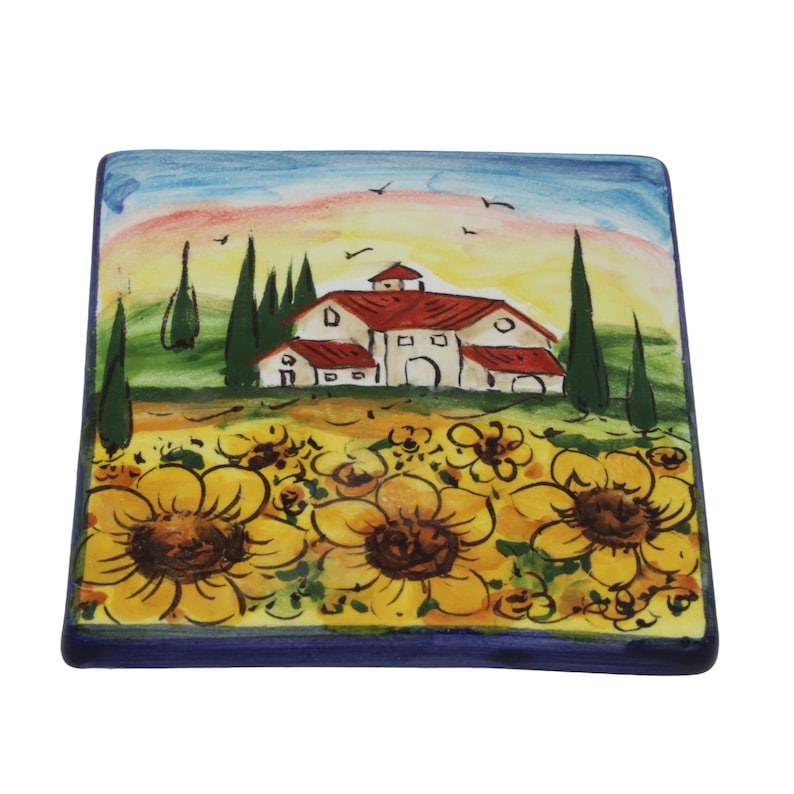 May include: Square ceramic coaster with a hand-painted design. The artwork depicts a rural scene with a farmhouse, sunflowers, and trees under a colorful sky. The coaster has a dark blue border.