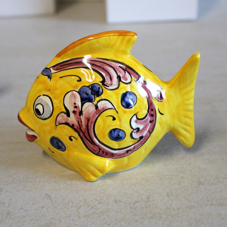 May include: A ceramic fish figurine in bright yellow with decorative painted details. The fish has a smiling mouth, a dorsal fin, and a tail fin. The body is adorned with burgundy, white, and blue floral patterns. The figurine is approximately 6 inches long.