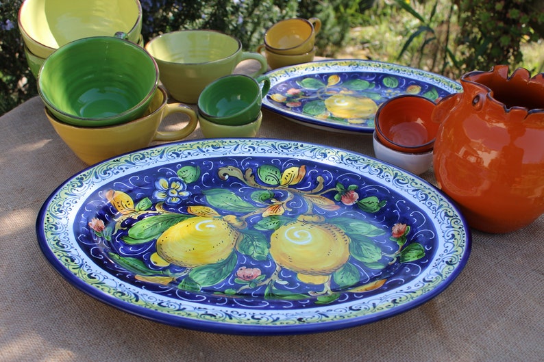 Puede incluir: Una colecci&oacute;n de vajilla de cer&aacute;mica colorida, que incluye fuentes, cuencos y tazas. La fuente ovalada presenta un fondo azul vibrante con un dise&ntilde;o de lim&oacute;n y flores. Otras piezas son amarillas, verdes y naranjas.
