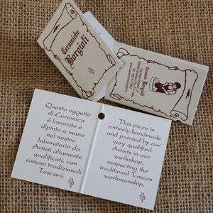 May include: Off-white product tags with burgundy text and illustrations. The tags read "Ceramiche Borgioli" and describe the handmade ceramic pieces, painted by qualified artists in a Tuscan workshop.