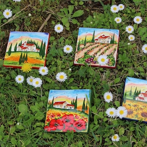 May include: Four square ceramic tiles with Italian landscape paintings, featuring houses, trees, and fields of flowers. The tiles are surrounded by green grass and white daisies.