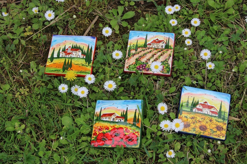 May include: Four square ceramic tiles with Italian landscape paintings, featuring houses, trees, and fields of flowers. The tiles are surrounded by green grass and white daisies.