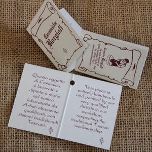 May include: Off-white product tags with the text "Ceramiche Borgioli" in brown script. The tags have a scroll design and state the products are handmade and painted by qualified artists in a Tuscan workshop.