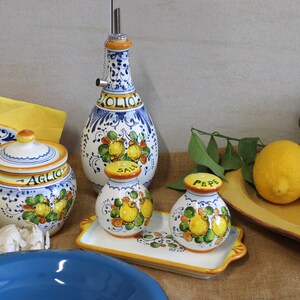 May include: A collection of Italian ceramic kitchenware featuring a blue and white design with lemon and leaf motifs. Includes an olive oil bottle, salt and pepper shakers, a garlic jar, and a serving tray. A lemon sits on a yellow tray.