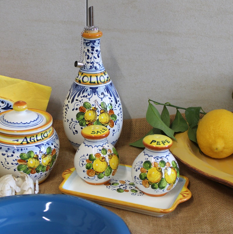 May include: A collection of Italian ceramic kitchenware featuring a blue and white design with lemon and leaf motifs. Includes an olive oil bottle, salt and pepper shakers, a garlic jar, and a serving tray. A lemon sits on a yellow tray.