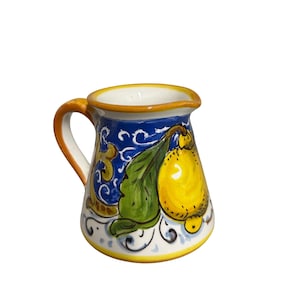 Puede incluir: Una jarrita de cerámica para crema con un limón amarillo pintado a mano y una hoja verde sobre un fondo azul vibrante. La jarra tiene un asa curva, un borde amarillo y patrones decorativos de volutas blancas alrededor de la base.