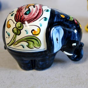 May include: A ceramic elephant figurine with a dark blue body and a white section decorated with a floral design in red, green, and yellow. The elephant's ear is light blue. This decorative item is a unique home decor piece.