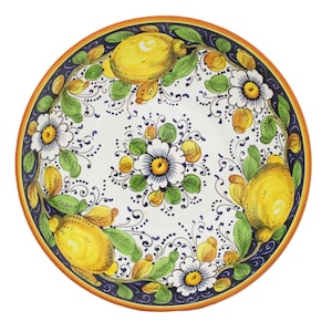 Può includere: Un piatto rotondo in ceramica con un disegno dipinto a mano. Il piatto presenta uno sfondo bianco con limoni gialli, foglie verdi e fiori bianchi. Un bordo blu scuro con un bordo arancione circonda il disegno. Il piatto è probabilmente per servire cibo.