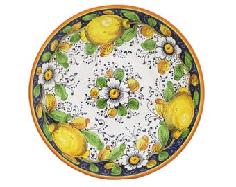 May include: A round ceramic plate with a hand-painted design. The plate features a white background with yellow lemons, green leaves, and white flowers. A dark blue border with an orange rim surrounds the design. The plate is likely for serving food.