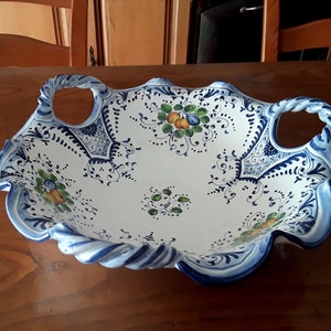 K&ouml;nnte beinhalten: Wei&szlig;es Keramik-Serviertablett mit blau-wei&szlig;en Blumenmustern und gedrehten Griffen. Die Schale hat einen gewellten Rand und ist mit gr&uuml;nen, gelben und blauen Obst- und Blumendesigns verziert. Ein dekoratives St&uuml;ck zum Servieren.