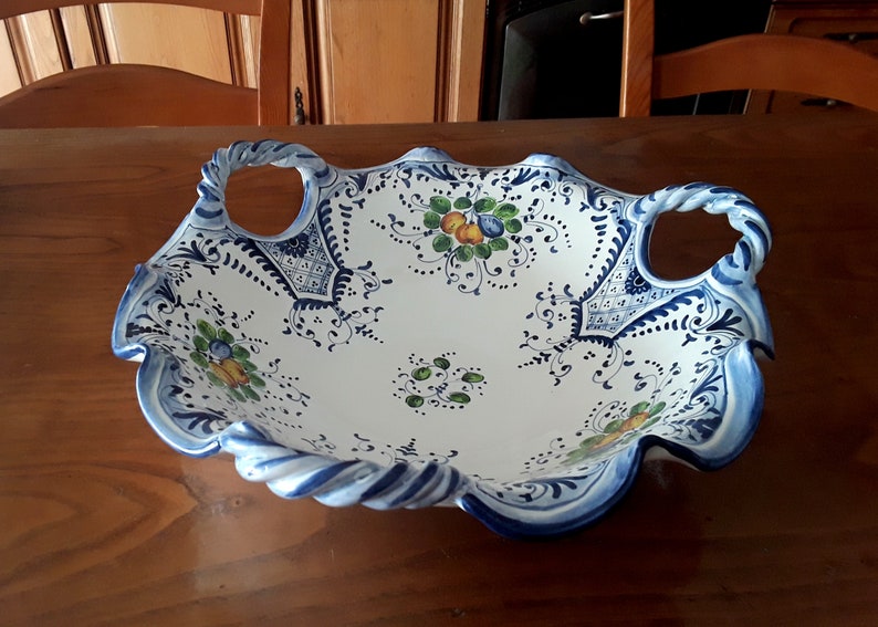 K&ouml;nnte beinhalten: Wei&szlig;es Keramik-Serviertablett mit blau-wei&szlig;en Blumenmustern und gedrehten Griffen. Die Schale hat einen gewellten Rand und ist mit gr&uuml;nen, gelben und blauen Obst- und Blumendesigns verziert. Ein dekoratives St&uuml;ck zum Servieren.