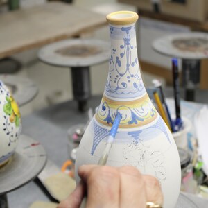 May include: A ceramic vase being hand-painted with blue and yellow floral designs. The vase has a narrow neck and a wide base. The artist is using a paintbrush to apply the paint. The vase is on a work surface.
