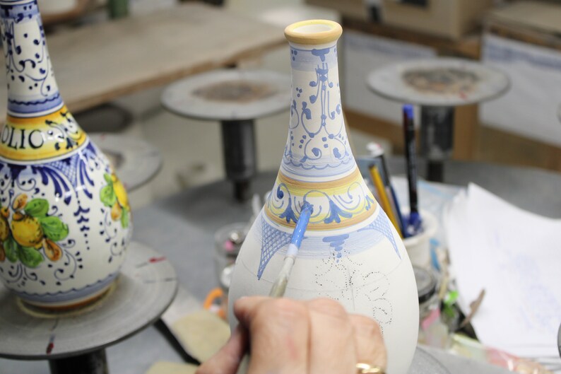 May include: A ceramic vase being hand-painted with blue and yellow floral designs. The vase has a narrow neck and a wide base. The artist is using a paintbrush to apply the paint. The vase is on a work surface.
