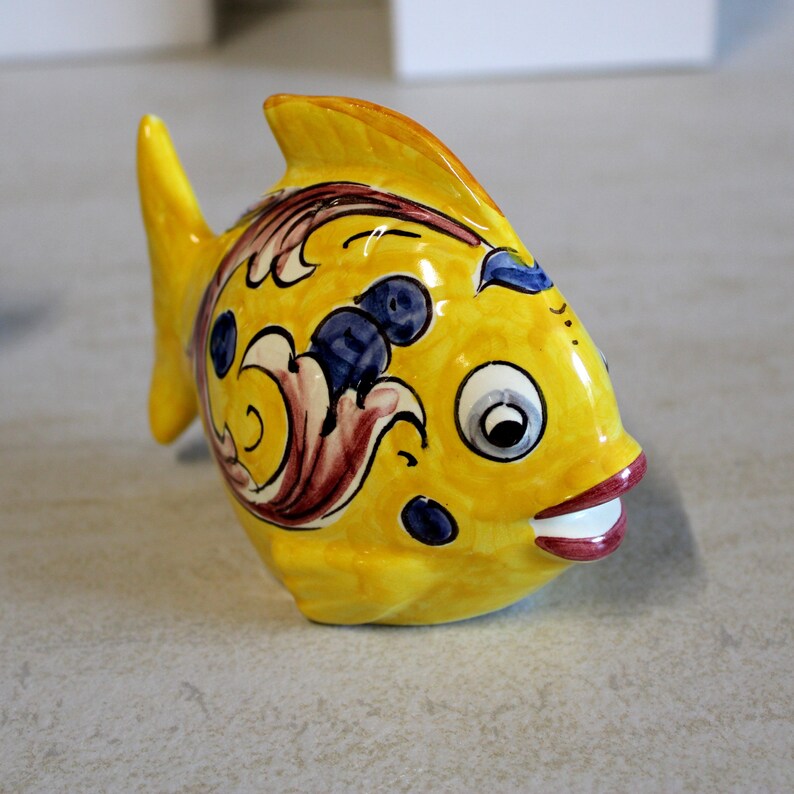 May include: A ceramic fish figurine in bright yellow with decorative blue, red, and white accents. The fish has a smiling mouth and large, expressive eyes. This decorative item is a whimsical addition to any home decor.