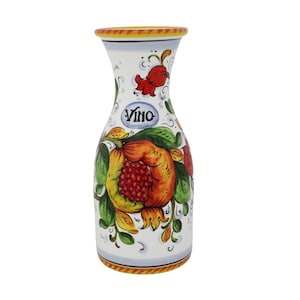 Può includere: Caraffa da vino in ceramica bianca con un colorato motivo floreale. La caraffa presenta un grande melograno e la scritta "Vino" in un ovale blu. Il bordo è decorato con accenti gialli e arancioni.