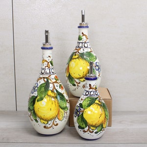 Può includere: Tre bottiglie di olio d'oliva in ceramica bianca con un design a limone. Ogni bottiglia ha un beccuccio argentato e la scritta "OLIO" stampata sopra. Le bottiglie sono decorate con limoni gialli, foglie verdi e fiori blu.