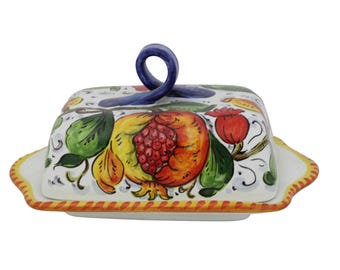 May include: A ceramic butter dish with a lid and a handle. The dish is white with a colorful hand-painted design of pomegranates, flowers, and leaves. The rim is decorated with a yellow and red pattern. The handle is blue.