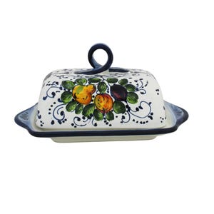 Può includere: Una burriera in ceramica bianca con coperchio, con un disegno dipinto a mano di frutta gialla e arancione con foglie verdi. Il coperchio ha un manico blu scuro. Il piatto ha un bordo e una base blu scuro.