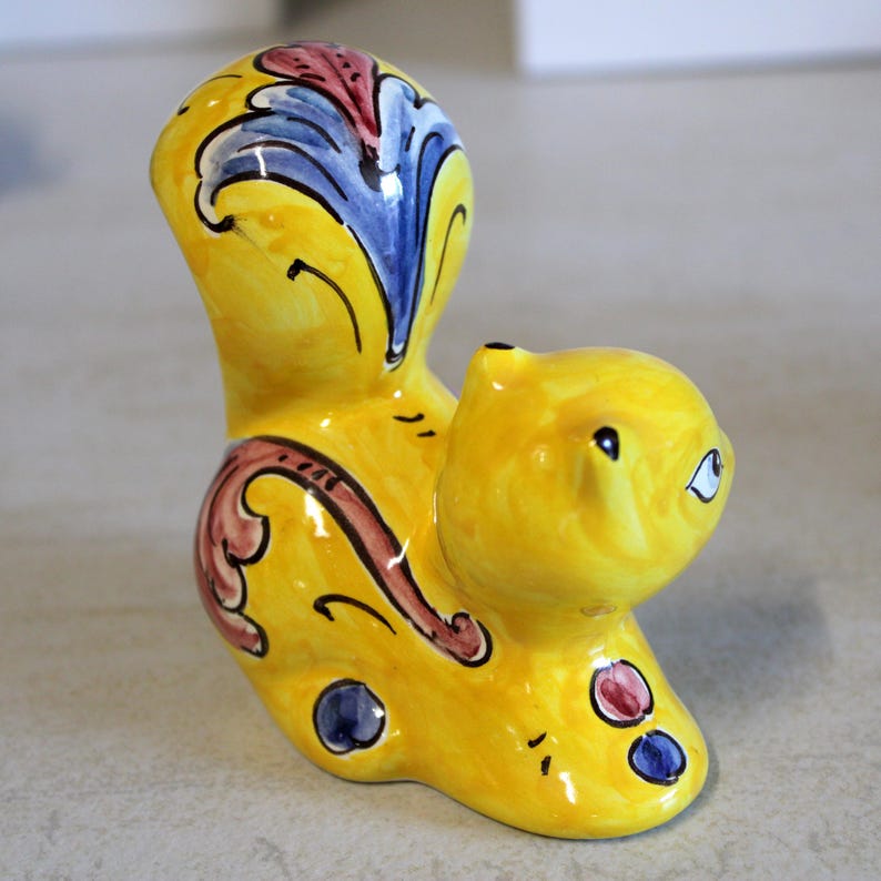May include: A ceramic squirrel figurine in bright yellow with decorative accents. The squirrel has a large, curved tail and is adorned with blue, red, and brown floral patterns. The figurine has a glossy finish and is a decorative item.