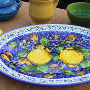 Puede incluir: Plato ovalado de cer&aacute;mica con un vibrante fondo azul, adornado con limones pintados a mano, hojas verdes y detalles florales. El borde presenta un borde decorativo blanco y amarillo. Otras piezas de cer&aacute;mica en el fondo.