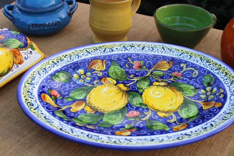 Puede incluir: Plato ovalado de cer&aacute;mica con un vibrante fondo azul, adornado con limones pintados a mano, hojas verdes y detalles florales. El borde presenta un borde decorativo blanco y amarillo. Otras piezas de cer&aacute;mica en el fondo.