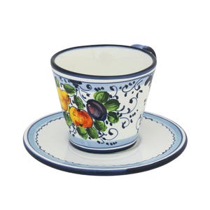 May include: A ceramic teacup and saucer set. The cup features a white background with a blue rim and handle, and is decorated with colorful fruit and floral designs. The saucer has a matching blue rim and a white center.