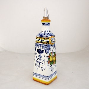 Può includere: Una bottiglia di olio d'oliva in ceramica bianca con un design floreale blu e giallo. La bottiglia ha un tappo di sughero e un beccuccio d'argento con una catena. La bottiglia è etichettata "Olio" in scrittura dorata.