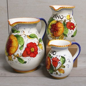 Puede incluir: Tres jarras de cerámica con ribetes azules y amarillos, decoradas con flores rojas y granadas. La jarra más grande dice "Vino" en letras rojas.