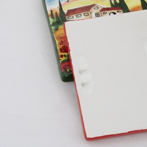 May include: Two rectangular ceramic tiles. One tile features a painted landscape with houses, trees, and a sunset. The other tile is white with a red border and has handwritten text. The tiles are on a white surface.