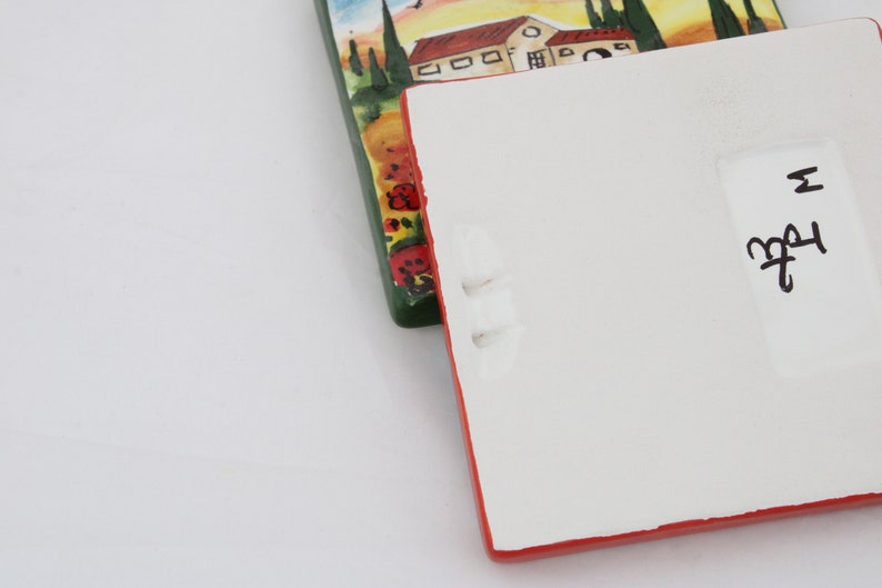May include: Two rectangular ceramic tiles. One tile features a painted landscape with houses, trees, and a sunset. The other tile is white with a red border and has handwritten text. The tiles are on a white surface.