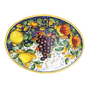 May include: Oval ceramic serving platter with a hand-painted design. The platter features a dark blue border with yellow and green foliage, lemons, pomegranates, and a bunch of grapes. The rim is trimmed with a gold-colored band.
