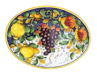 May include: Oval ceramic serving platter with a hand-painted design. The platter features a dark blue border with yellow and green foliage, lemons, pomegranates, and a bunch of grapes. The rim is trimmed with a gold-colored band.