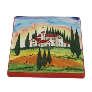May include: Square ceramic tile featuring a hand-painted Italian landscape scene. The artwork depicts a white building with a red roof, surrounded by green trees and fields, under a colorful sunset sky. The tile has a red border.