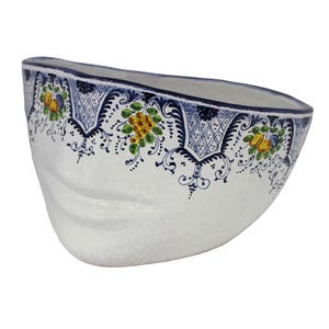 Può includere: Ciotola in ceramica bianca con un motivo floreale blu e giallo sul bordo. La ciotola ha una forma asimmetrica unica e una superficie strutturata. Il bordo è dipinto con un bordo blu scuro.